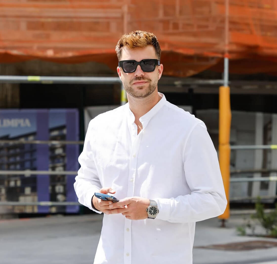 Hombre con camisa blanca y gafas de sol negras sostiene un teléfono móvil al aire libre.