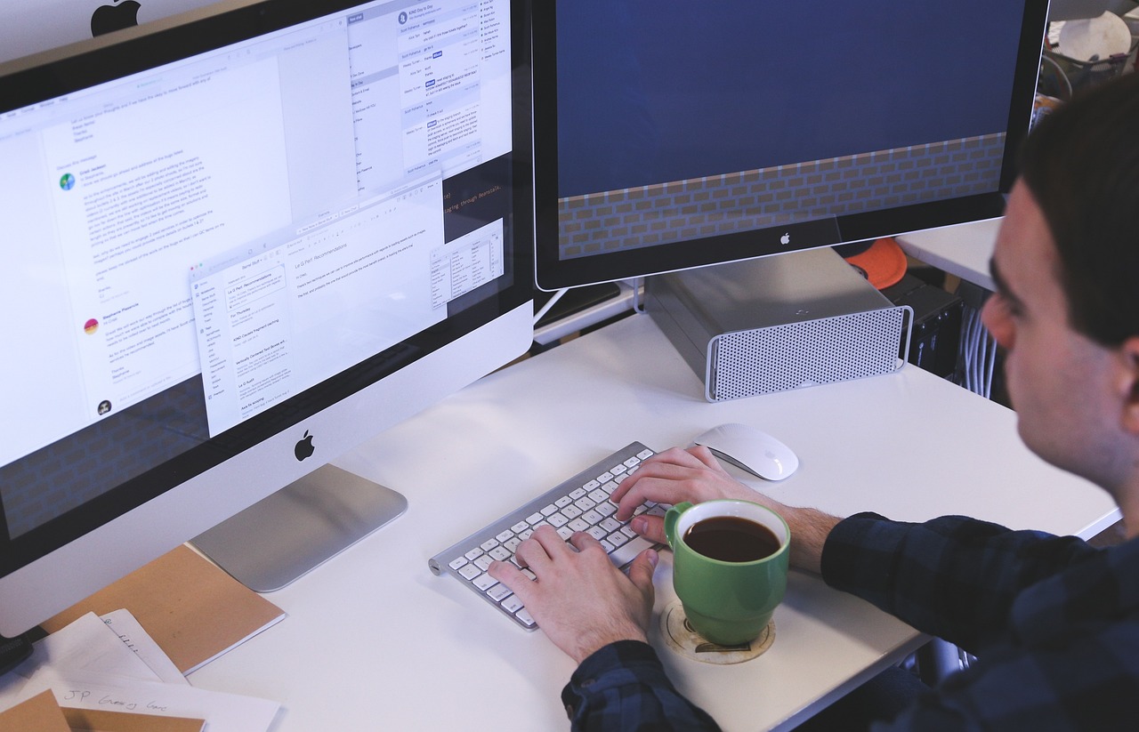 Persona escribiendo en un teclado inalámbrico frente a dos monitores Apple en un escritorio blanco con una taza verde de café.